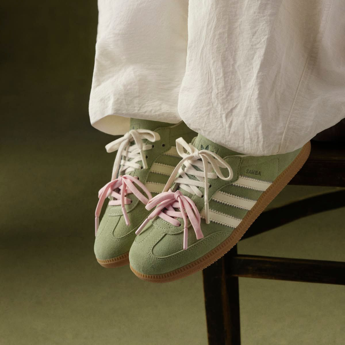 Green Adidas sneakers with pink laces worn by a person sitting on a chair.