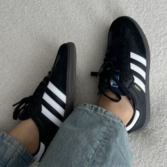Black Adidas sneakers with white stripes worn with light blue jeans on a neutral background