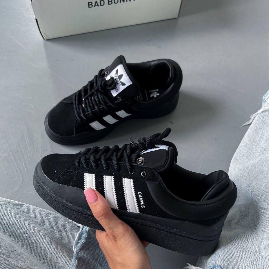 Black Adidas sneakers with white stripes held by a hand on a light gray surface.