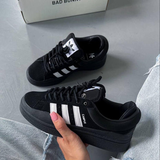 Black Adidas sneakers with white stripes held by a hand on a light gray surface.