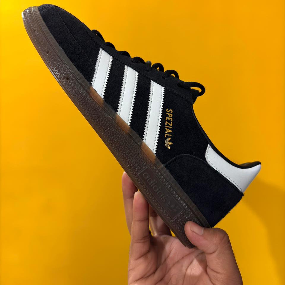 Black Adidas sneaker with white stripes held against a yellow background