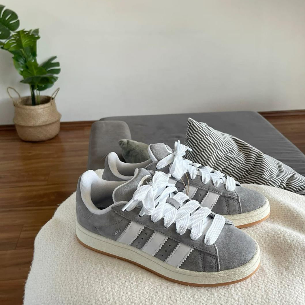 Gray Adidas sneakers with white laces on a beige surface, with a plant and gray cushion in the background.