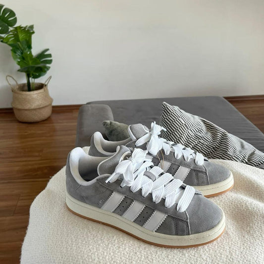 Gray Adidas sneakers with white laces on a beige surface, with a plant and gray cushion in the background.