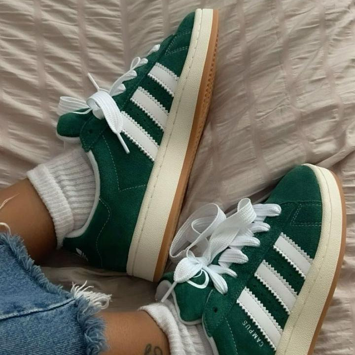 Green Adidas sneakers with white stripes worn indoors on a wooden floor.