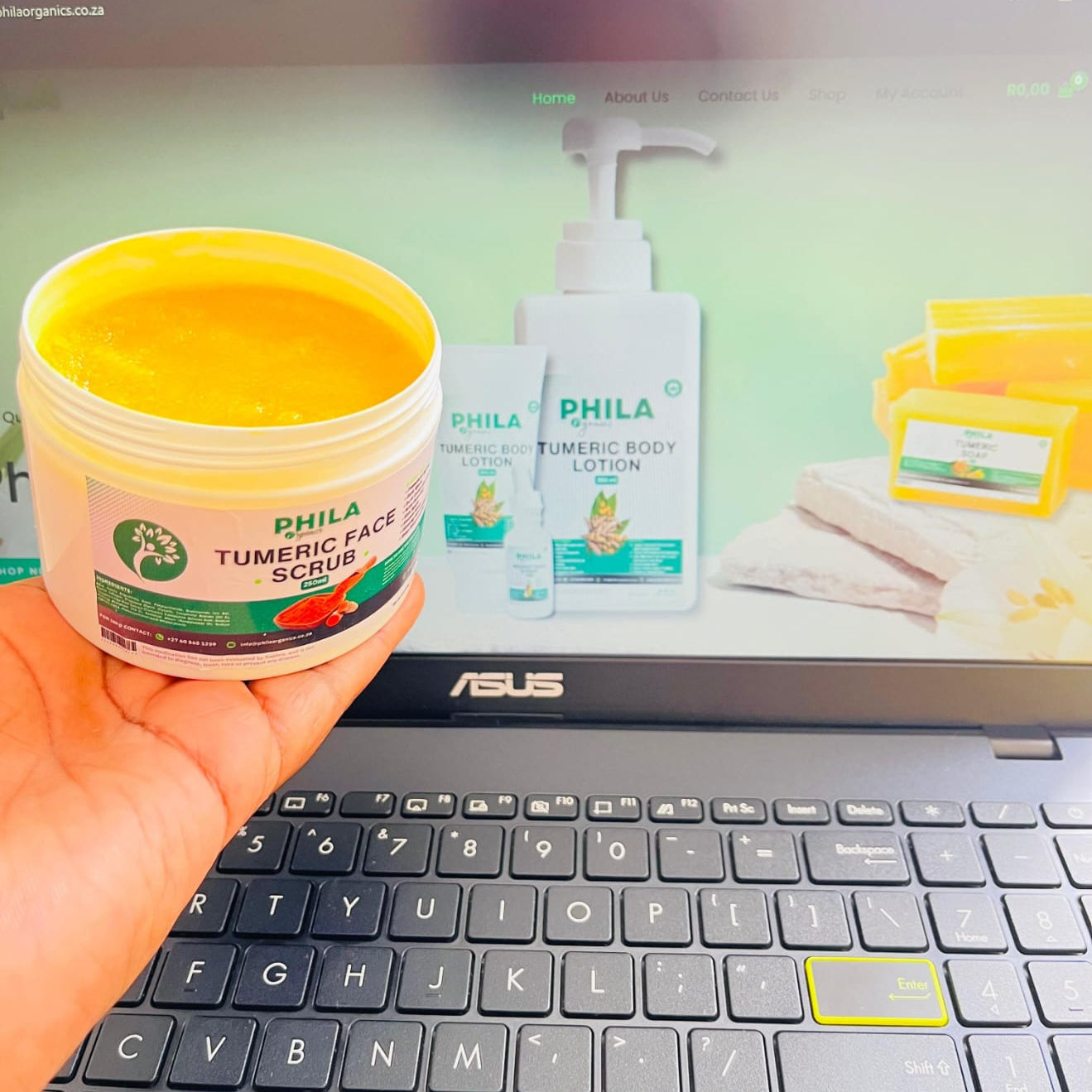 Person holding a jar of PHILA Tumeric Face Scrub in front of an ASUS laptop displaying PHILA products.