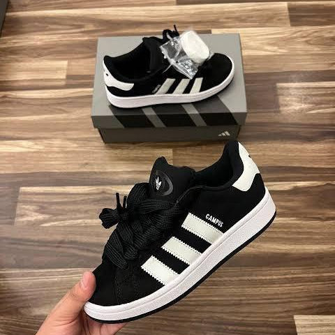 Black and white Adidas sneakers held by a hand on a wooden floor with a box in the background.