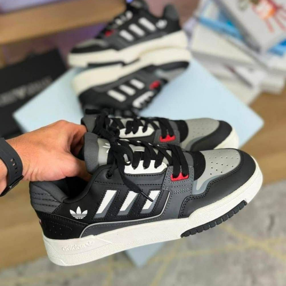 Person holding a black and gray Adidas sneaker with white soles on a blurred indoor background.