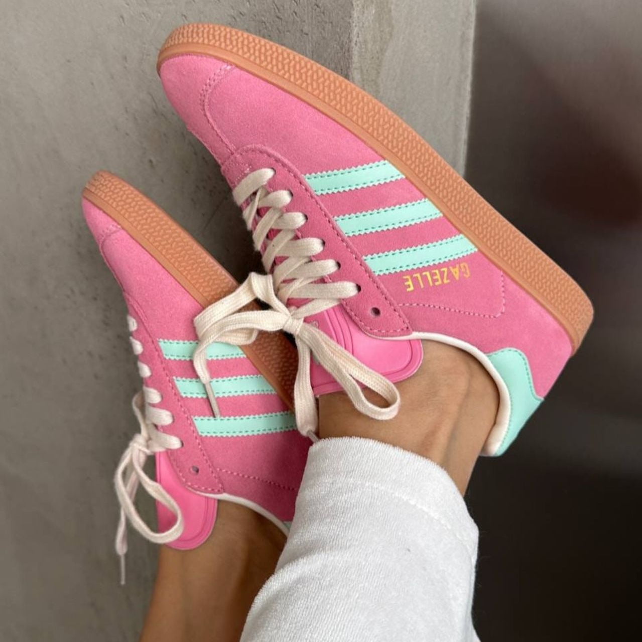Pink Adidas sneakers with turquoise stripes worn by a person against a neutral background