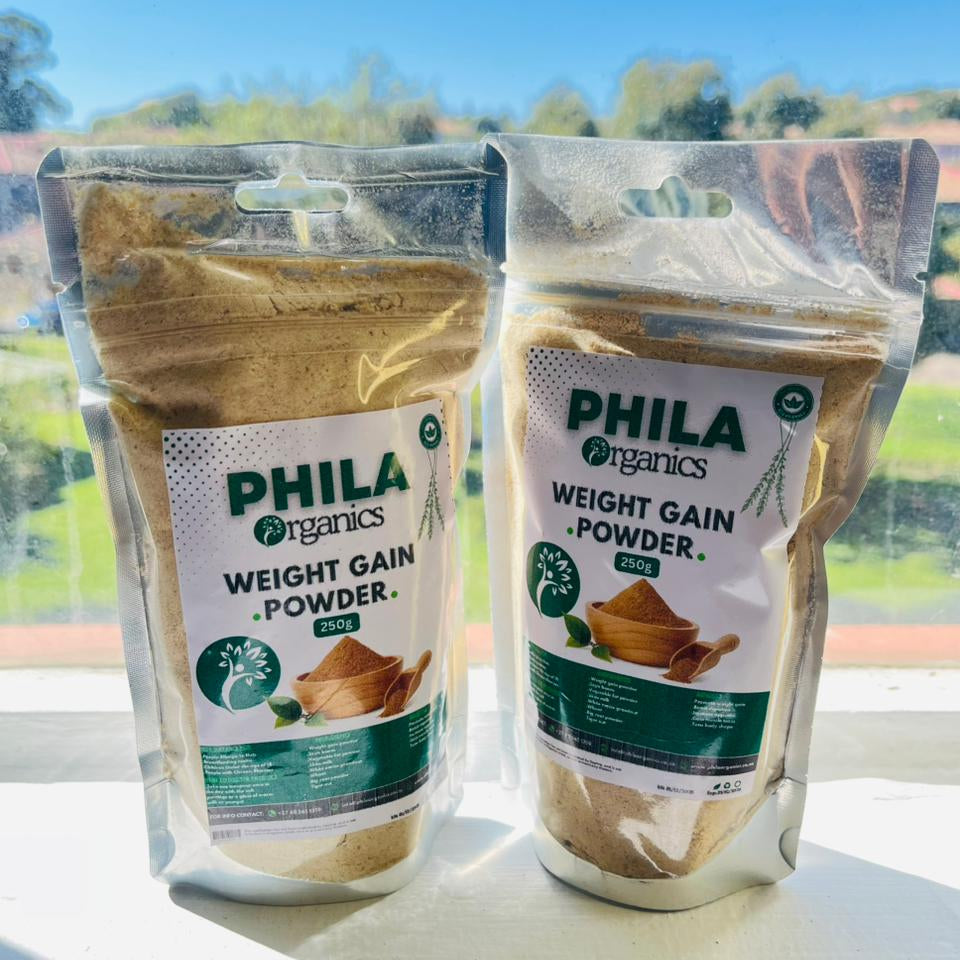 Two packages of Phila Organics weight gain powder on a windowsill with a view of trees outside.