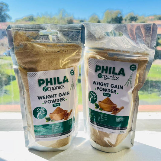 Two packages of Phila Organics weight gain powder on a windowsill with a view of trees outside.